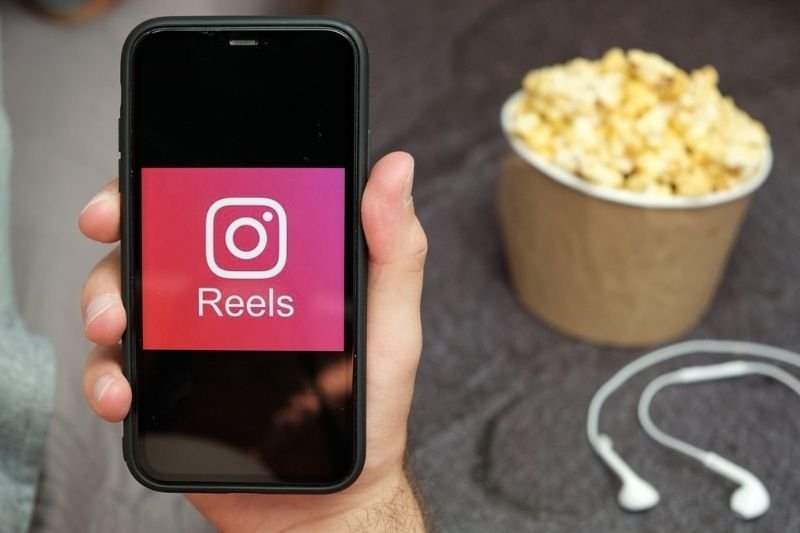 A hand holds a smartphone displaying the Instagram Reels logo, with popcorn and earphones nearby.