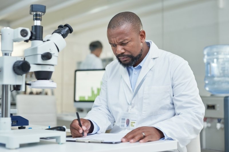 A male medical researcher in a white lab coat reviews documents beside a microscope in a laboratory.