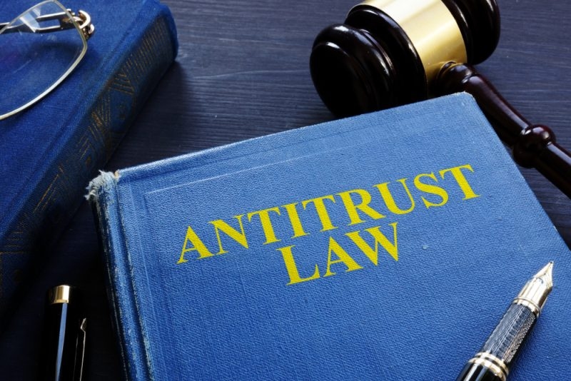 Blue law book labeled “ANTITRUST LAW” beside a judge’s gavel, eyeglasses, and fountain pen on a dark wooden surface.
