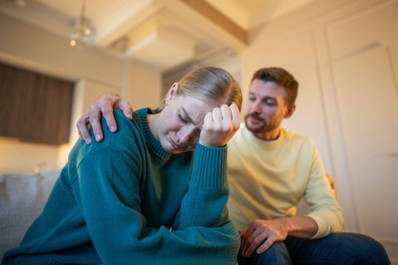 Woman appearing distressed while a man offers comfort by placing a hand on her shoulder at home.