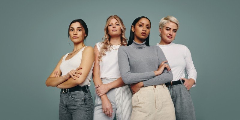 Four women of different appearances and styles stand together against a muted green background, wearing casual contemporary outfits.