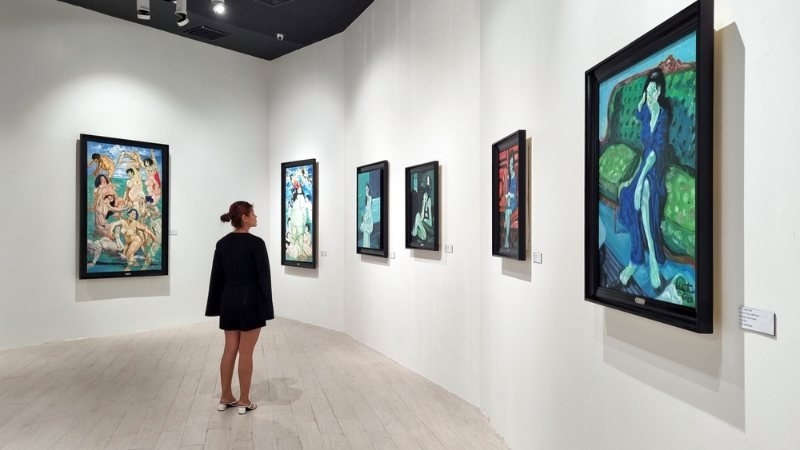 A visitor stands in a gallery surrounded by colorful framed paintings displayed on white walls.