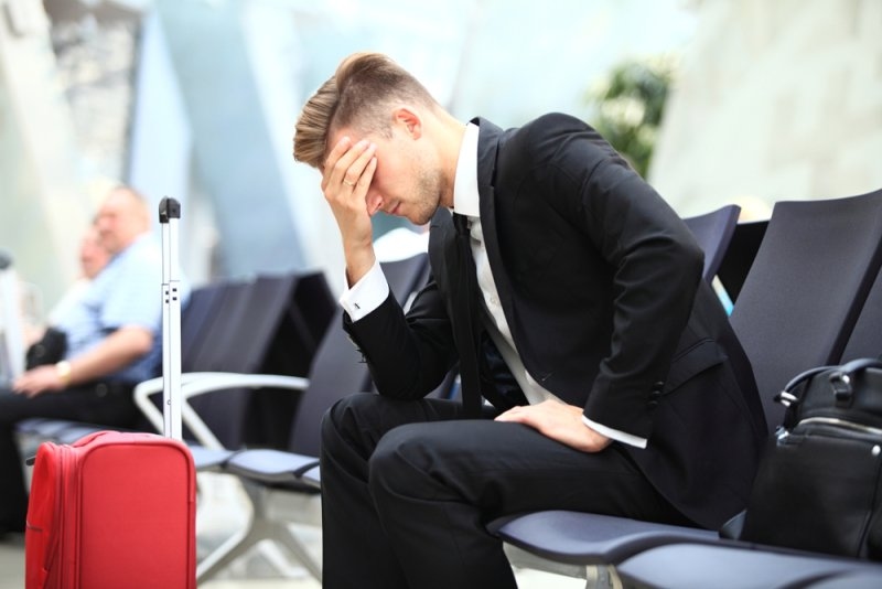 Frustrated businessman sitting in an airport waiting area beside his red suitcase.