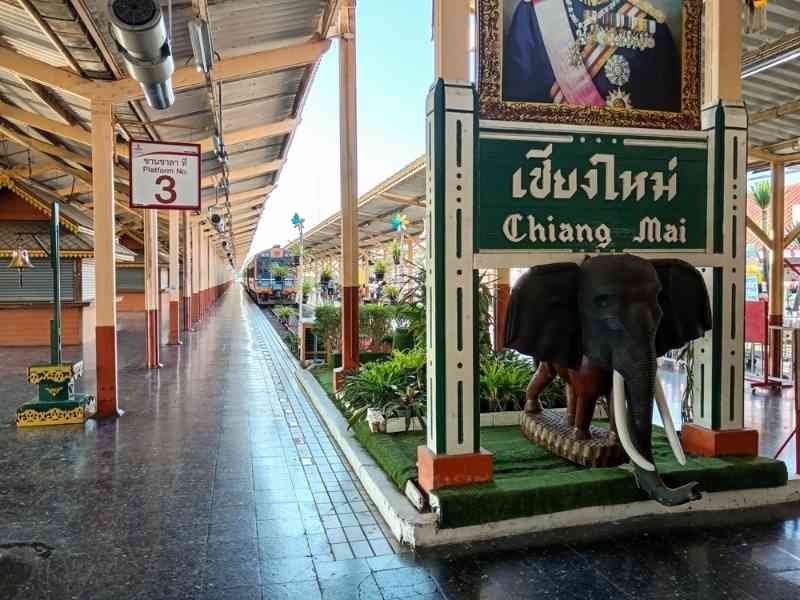 A train station platform in Chiang Mai, Thailand, featuring a bilingual station sign and a decorative elephant statue near the entrance.