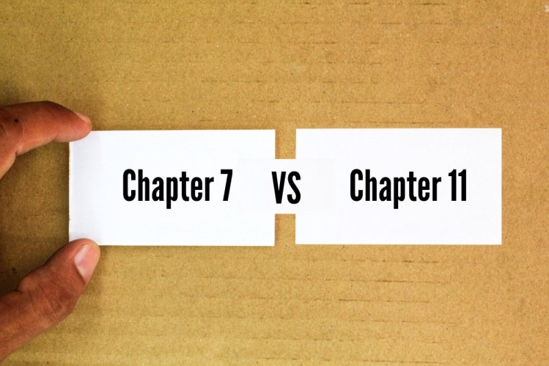 Two cards comparing “Chapter 7” and “Chapter 11” bankruptcy, held over a wooden surface.