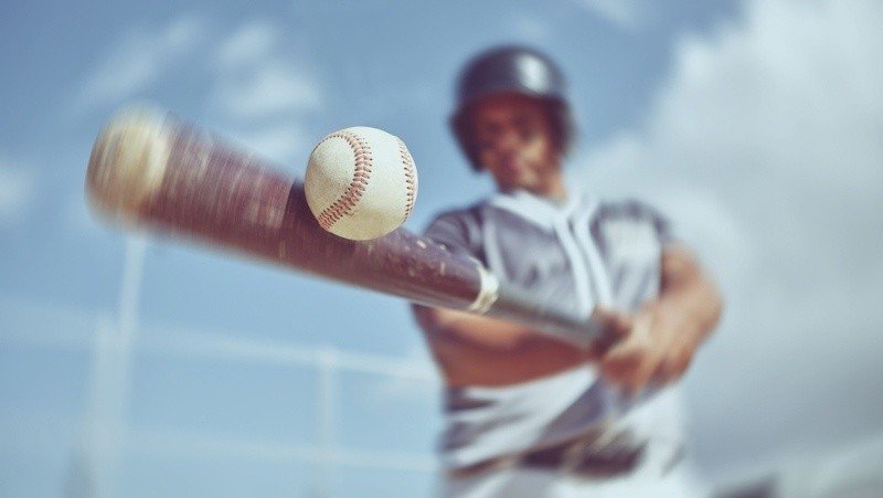 A baseball player swings a bat toward an approaching baseball on a bright outdoor field.
