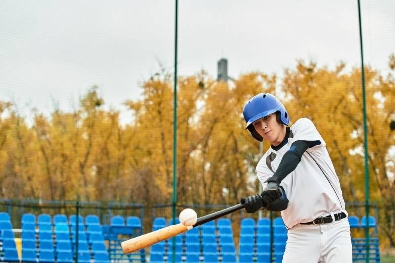 Young baseball player hitting a ball on a field with autumn trees in the background.