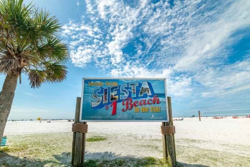 A sign for Siesta Beach in Sarasota, Florida stands on a sunny white-sand beach.