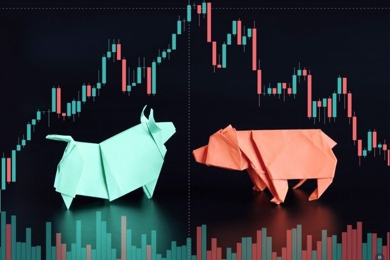 Paper origami bull and bear figures placed in front of a candlestick trading chart.