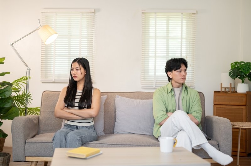 Couple sitting apart on a sofa with crossed arms and tense expressions, suggesting relationship conflict or emotional distance.