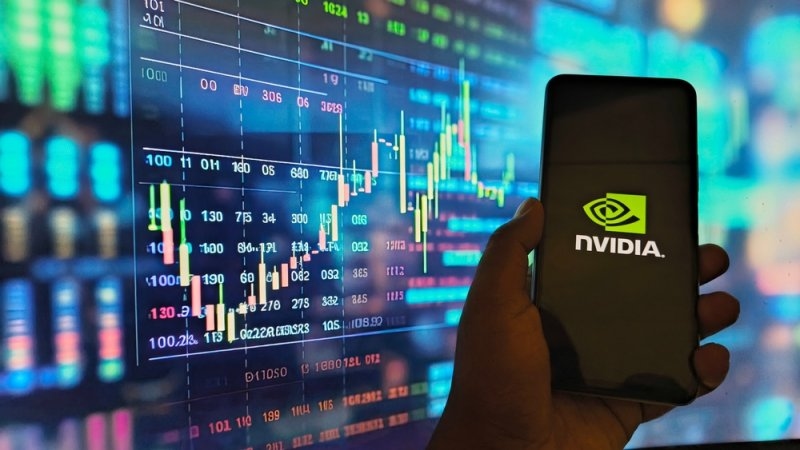 Hand holding a smartphone displaying the NVIDIA logo in front of a digital stock market chart with financial data and candlestick graphs.