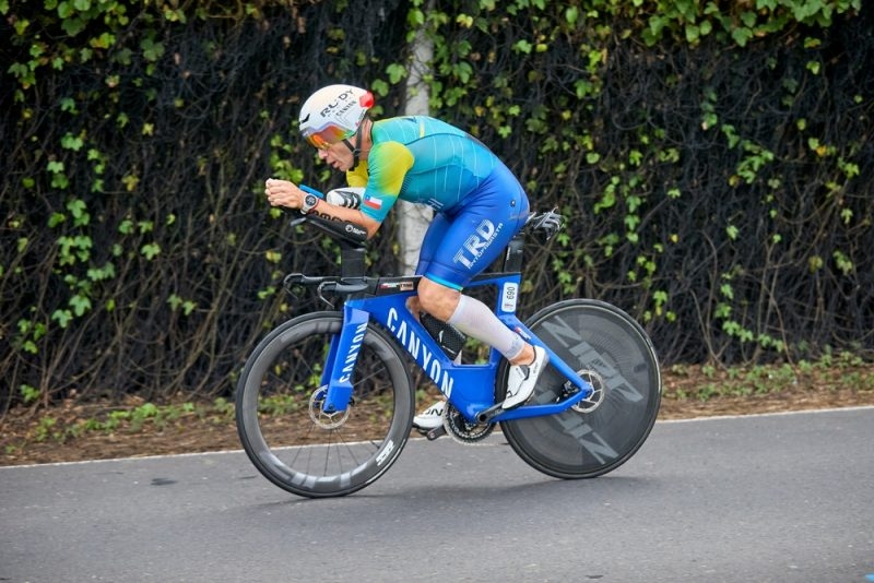 Cyclist wearing a blue and green racing kit riding an aerodynamic road bike on a paved road, with trees in the background.