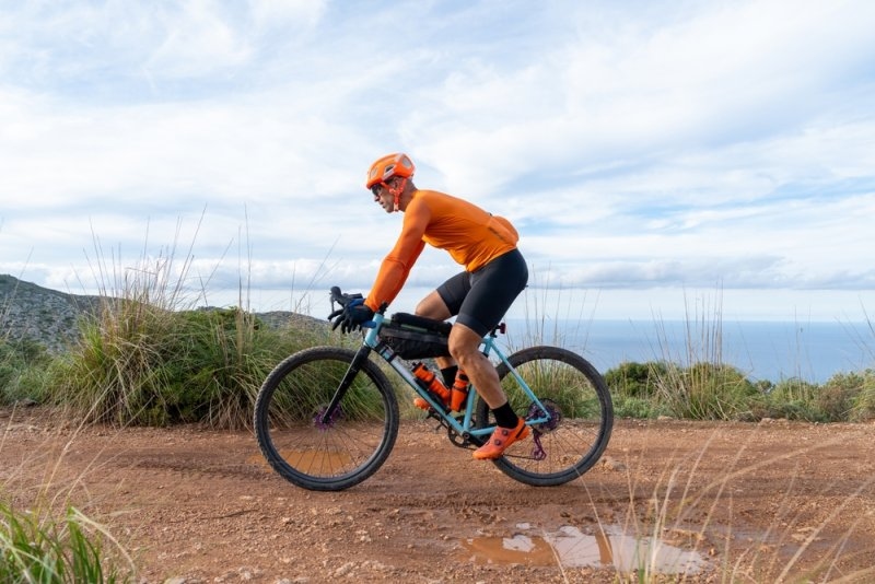 Cyclist wearing an orange helmet and matching jersey riding a gravel bike along a dirt trail with a coastal landscape in the background.