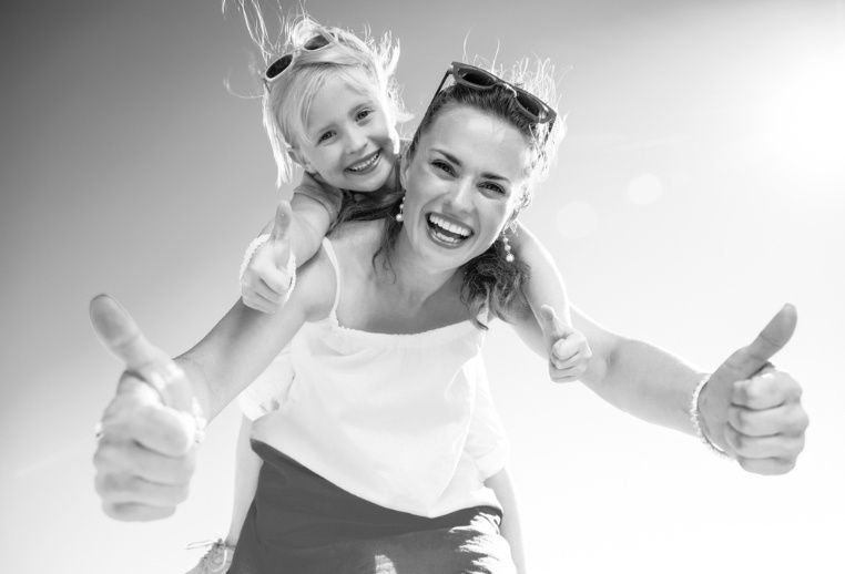 A smiling mother carries a young child on her shoulders, with both giving thumbs-up gestures in a bright black-and-white photograph.