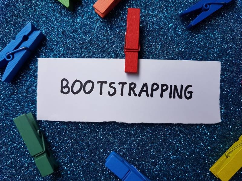 White card labeled “BOOTSTRAPPING” clipped to a colorful clothespin arrangement on a blue textured background.