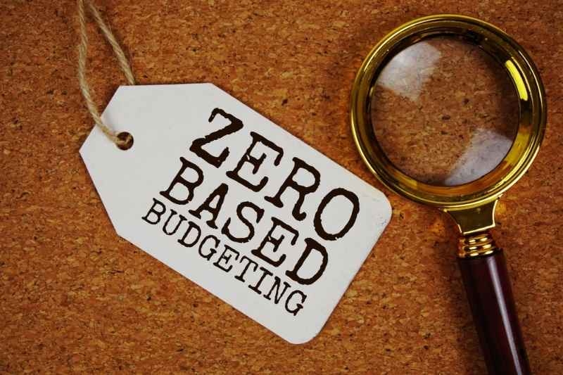 Tag labeled 'Zero Based Budgeting' beside a magnifying glass on a corkboard background.