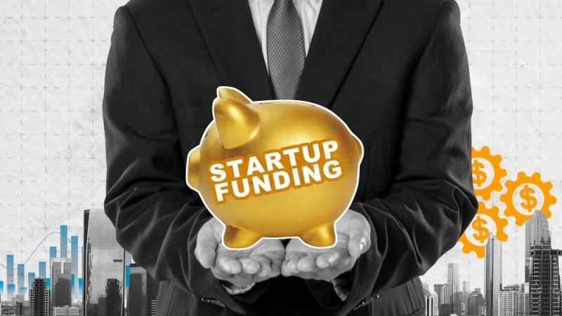 A businessman holding a piggy bank labeled Startup Funding against a city background with financial charts and dollar sign gears.