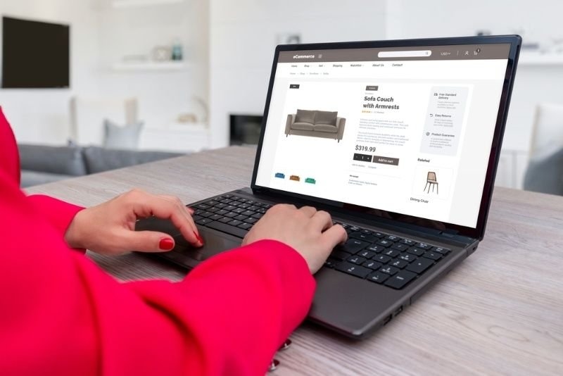A person uses a laptop to browse an e-commerce website featuring a sofa with armrests.
