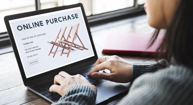 A person shopping online on a laptop, with the screen displaying an “ONLINE PURCHASE” advertisement for outdoor folding chairs.