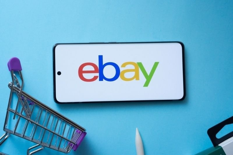 Smartphone displaying the eBay logo beside a miniature shopping cart, symbolizing online marketplace shopping.