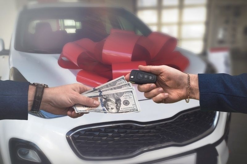 Person handing over cash while receiving a car key in front of a new car with a red bow.