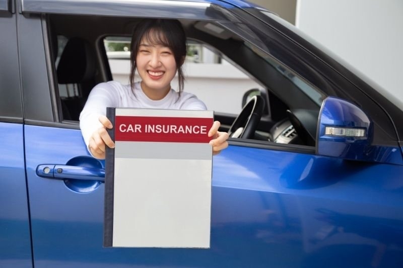 Smiling driver sitting in a blue car holding a document labeled "Car Insurance".