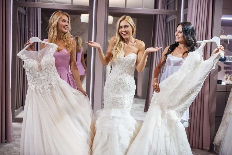 Bride standing between two friends in a bridal boutique while comparing wedding dresses and smiling during the dress selection process.