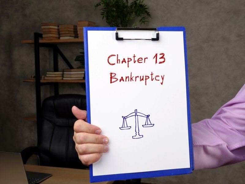 Person holding a clipboard labeled “Chapter 13 Bankruptcy” with a scales of justice symbol.