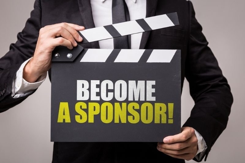 A person in a black suit holding a film clapperboard with the bold message “BECOME A SPONSOR!”.