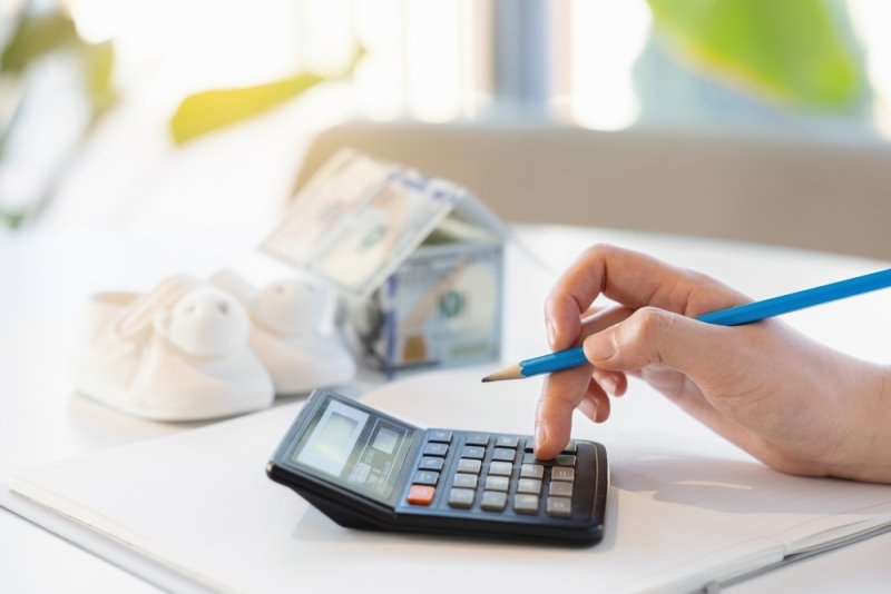 Person using a calculator to review financial expenses with money and baby shoes in the background.