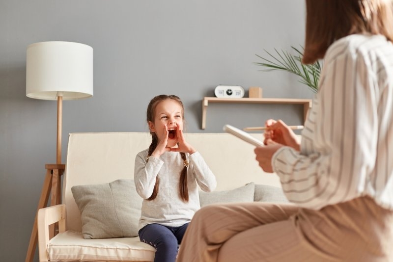 Little girl at therapy session with psychologist expressing her emotions and frustrations by shouting loudly while specialist calmly listens and takes notes.