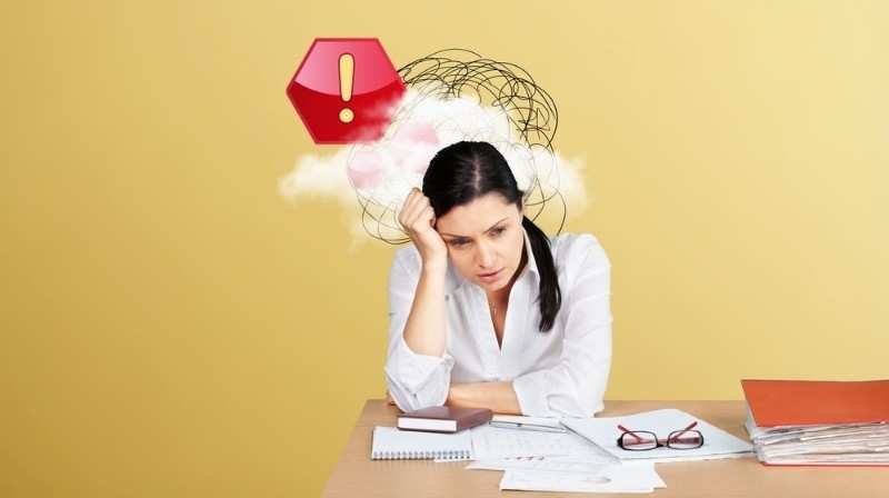 Stressed businesswoman sitting at a desk with papers and illustrated confusion symbols above her head.