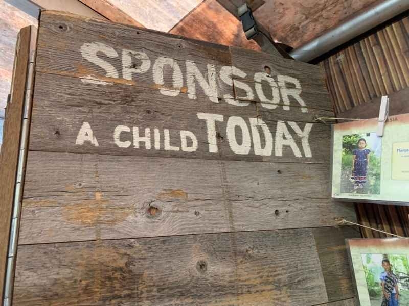 Photograph of a rustic wooden wall displaying the message “SPONSOR A CHILD TODAY,” with informational displays featuring photographs of children visible on the right.
