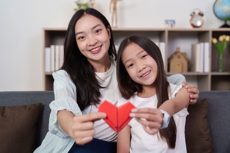 Mother and daughter smiling together while holding a red paper heart at home.