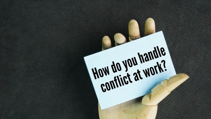 Wooden hand holding a card that asks, “How do you handle conflict at work?”