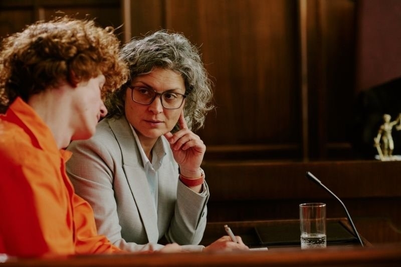 Middle aged Caucasian woman with gray hair and glasses listening attentively to teenage Caucasian boy with curly hair during legal consultation in courtroom setting.