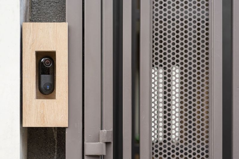 Modern smart video doorbell camera mounted on a wooden panel attached to a grey perforated metal gate for home security and surveillance