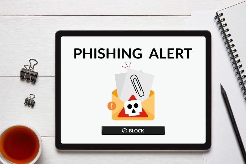 Overhead shot of a phishing alert displayed on a tablet screen, placed on a white wooden table surrounded by office items.