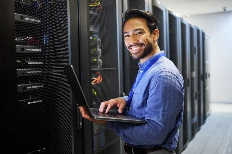 A smiling IT professional using a laptop in a server room while monitoring and maintaining data center equipment.