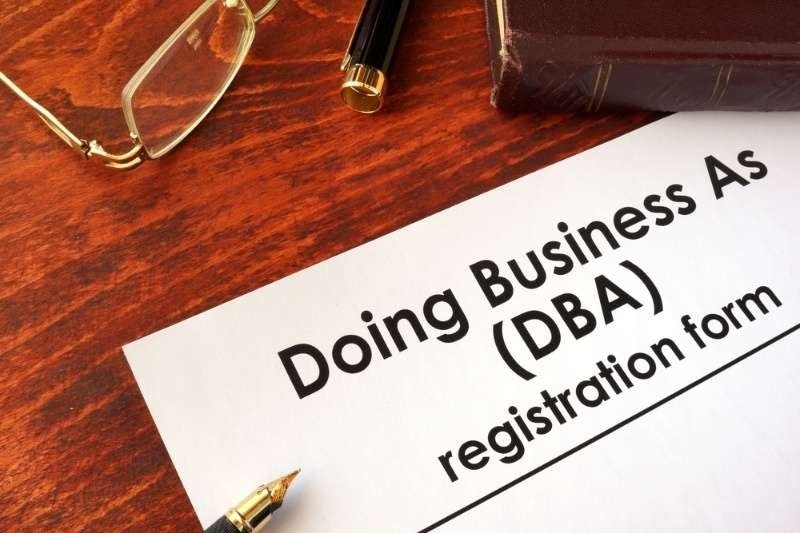 Close-up of a "Doing Business As (DBA) Registration Form" on a desk with eyeglasses, pen, and legal documents.