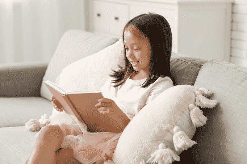Young girl sitting on a couch reading a book indoors, smiling while enjoying a quiet reading moment at home.