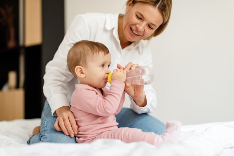 Mother helping a baby drink water from a bottle while sitting on a bed.
