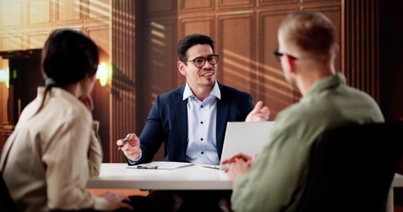 A business professional wearing glasses discusses documents with two colleagues across a table in an office.