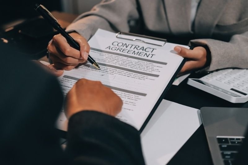 Two people are shown signing a document labeled “CONTRACT AGREEMENT” on a desk, with a calculator and laptop nearby.