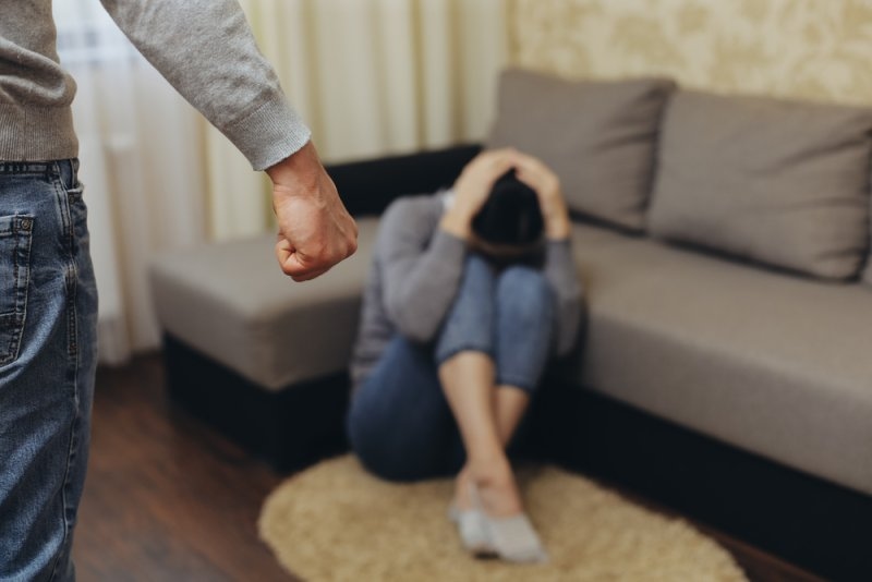 Person sitting distressed on the floor while another person stands nearby with a clenched fist, illustrating domestic violence.