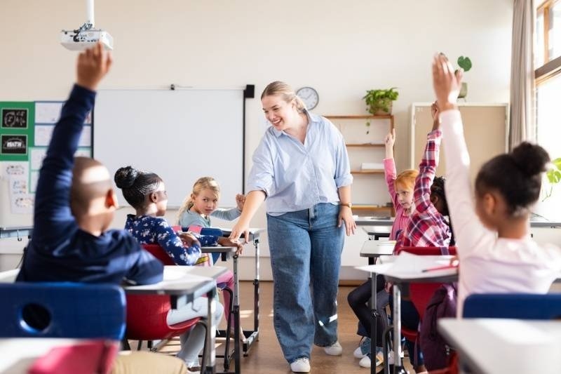 A teacher interacting with students in a bright classroom, with several children raising their hands to participate.