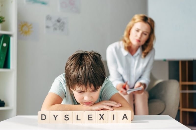 A child sitting at a desk with the word “DYSLEXIA” spelled out using wooden letter blocks, while an adult sits in the background.