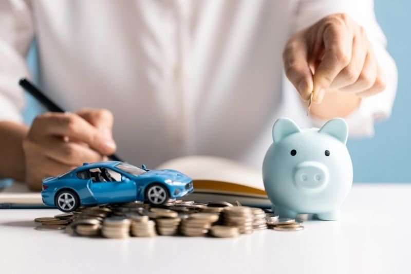 Person placing a coin into a piggy bank beside a model car and a stack of coins.