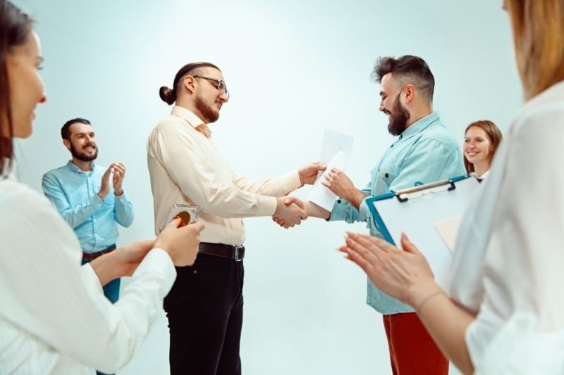 Manager presenting a certificate and shaking hands with an employee while coworkers applaud.