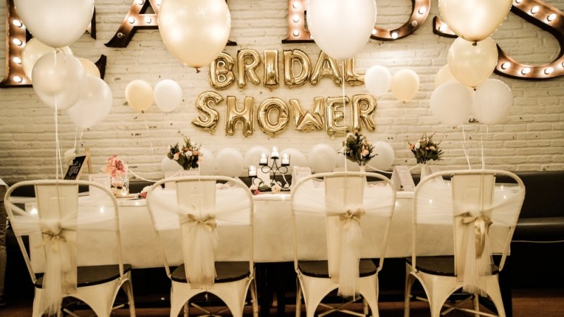 Decorated bridal shower venue with white balloons, gold “BRIDAL SHOWER” lettering, floral arrangements, and white-covered chairs.